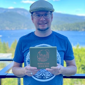 May include: A person wearing a blue t-shirt with a green box in their hands. The box has a white design of a globe with the text "Permaculture Design Deck" on it.