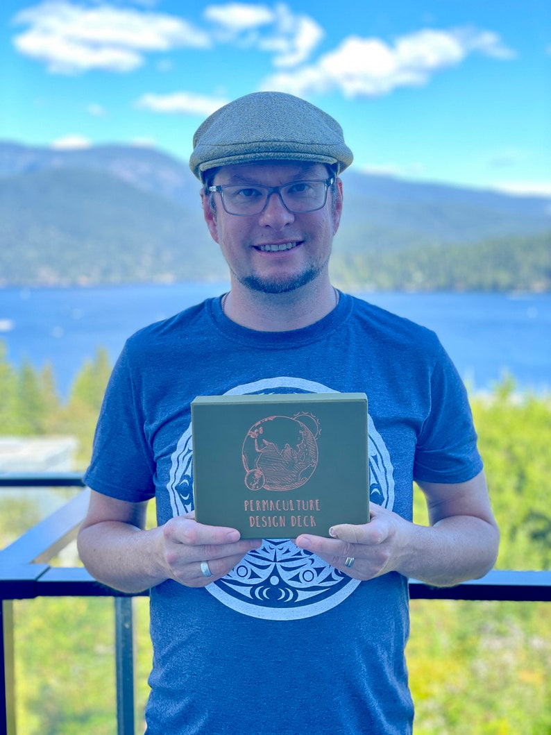 May include: A person wearing a blue t-shirt with a green box in their hands. The box has a white design of a globe with the text "Permaculture Design Deck" on it.