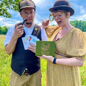 May include: Two people dressed in vintage clothing are holding a green box with the text "Permaculture Design Deck" on it. The woman is holding a magnifying glass and the man is holding a pipe and a deck of cards. They are standing in a field of tall grass.