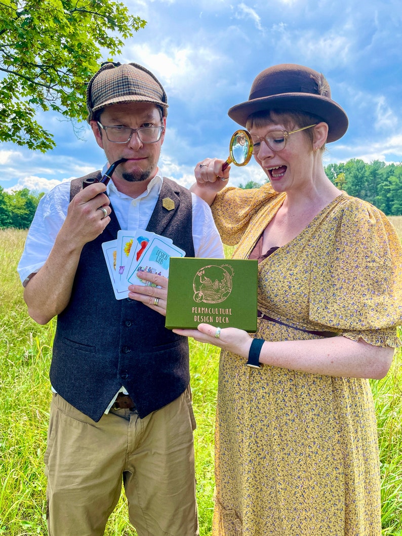 May include: Two people dressed in vintage clothing are holding a green box with the text "Permaculture Design Deck" on it. The woman is holding a magnifying glass and the man is holding a pipe and a deck of cards. They are standing in a field of tall grass.