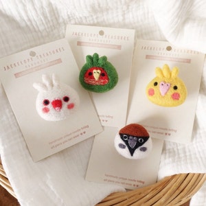 May include: Four felt animal pins on white cards with the text "Jackelope Treasures Handmade". The pins are a white cockatiel, a green and red parakeet, a yellow canary, and a brown and white sparrow.
