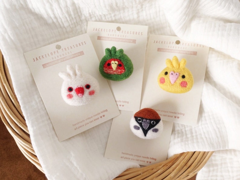May include: Four felt animal pins on white cards with the text "Jackelope Treasures Handmade". The pins are a white cockatiel, a green and red parakeet, a yellow canary, and a brown and white sparrow.