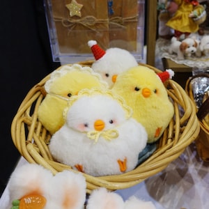 Puede incluir: Una cesta de mimbre llena de cuatro pollitos de peluche. Dos pollitos son amarillos con detalles blancos, uno es blanco con detalles amarillos y uno es blanco con un gorro de Papá Noel rojo.