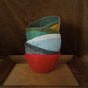 May include: A stack of five felt bowls in different colors, including red, green, blue, gray, and yellow. The bowls are nested inside each other and are sitting on a wooden surface.