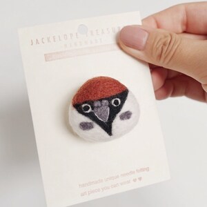 May include: A small, hand-felted bird pin with a brown head, white body, and black eyes. The pin is on a white card with the text "Jackelope Treasure Handmade" and "handmade unique needle felting art piece you can wear".
