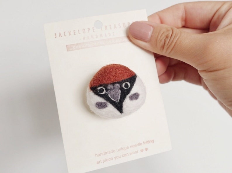 May include: A small, hand-felted bird pin with a brown head, white body, and black eyes. The pin is on a white card with the text "Jackelope Treasure Handmade" and "handmade unique needle felting art piece you can wear".