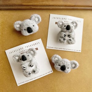 May include: Three needle felted koala brooches, each with a different pose. The brooches are gray and white with black eyes and noses. The brooches are displayed on a cork board with a white card behind each one. The card reads "Jackelope Treasures Handmade".