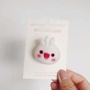 May include: White felt bunny pin with red cheeks and eyes. The pin is on a white card with the text "Jackelope Treasures Handmade" printed on it.