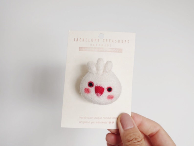 May include: White felt bunny pin with red cheeks and eyes. The pin is on a white card with the text "Jackelope Treasures Handmade" printed on it.