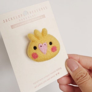 May include: A yellow felt needle felted cockatiel bird pin with pink accents. The pin is on a white card with the text "JACKELOPL TREASURES HARDMADE. CUSTOM BROOCHABLE" and "handmade unique needle felting art piece you can wear".