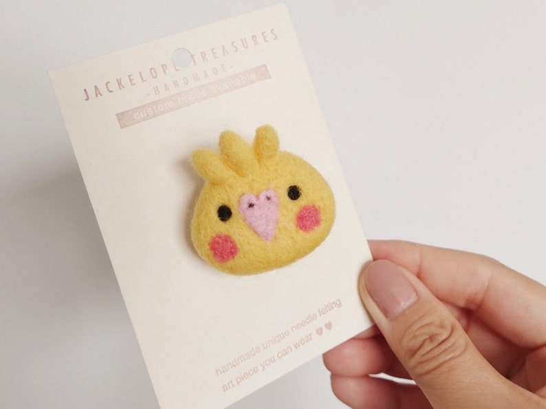 May include: A yellow felt needle felted cockatiel bird pin with pink accents. The pin is on a white card with the text "JACKELOPL TREASURES HARDMADE. CUSTOM BROOCHABLE" and "handmade unique needle felting art piece you can wear".