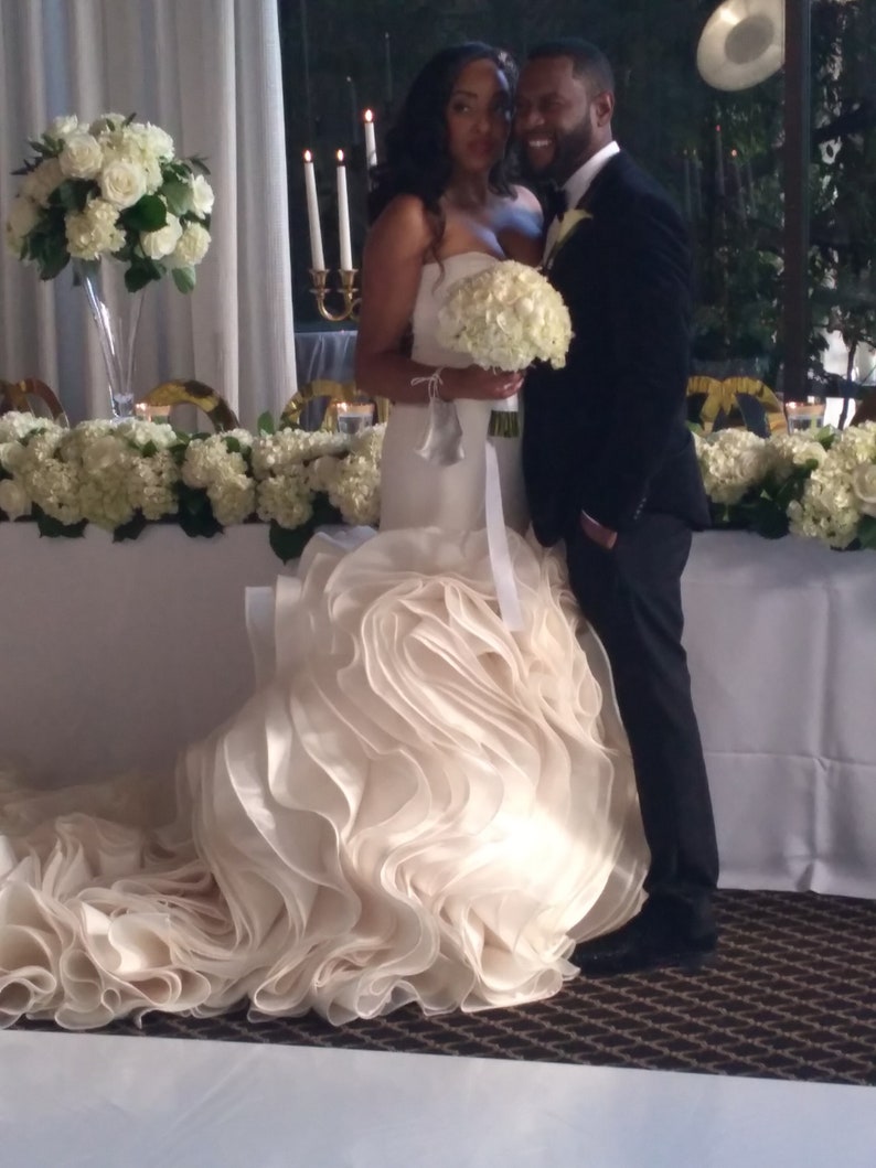 May include: A bride and groom pose for a wedding photograph. The bride is wearing a white strapless gown with a large, ruffled skirt. The groom is wearing a black suit and tie. They are standing in front of a white table with white flowers.