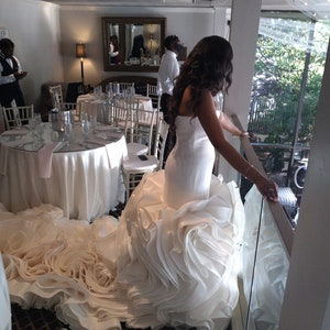 May include: A bride in a white wedding dress with a large, ruffled skirt stands on a balcony overlooking a room with tables and chairs. The dress has a fitted bodice and a long train.