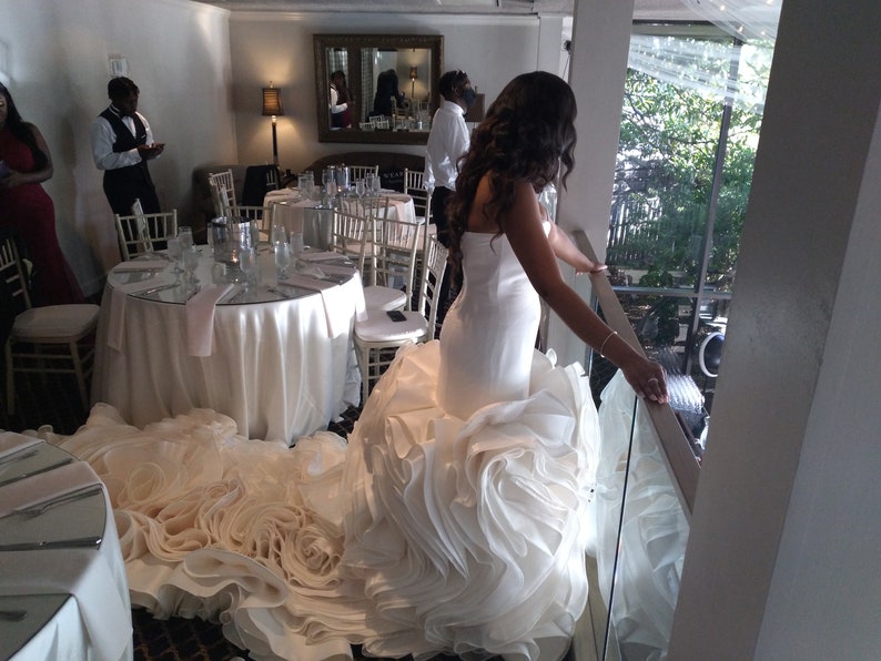 May include: A bride in a white wedding dress with a large, ruffled skirt stands on a balcony overlooking a room with tables and chairs. The dress has a fitted bodice and a long train.