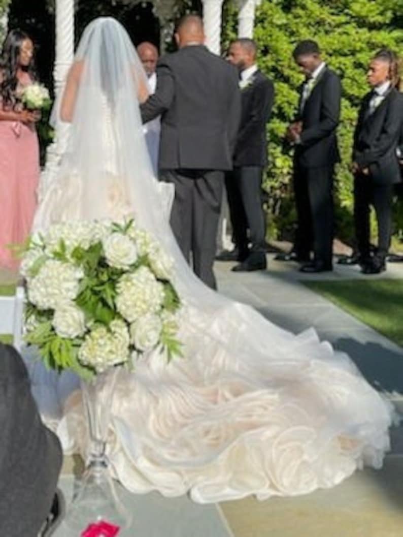 May include: A white wedding dress with a long train and a bouquet of white flowers. The dress has a fitted bodice and a full skirt.