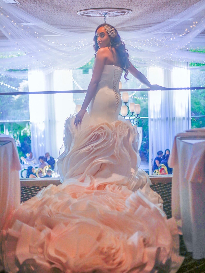 May include: A bride wearing a white strapless wedding dress with a large, ruffled skirt. The dress has a fitted bodice and a long train. The bride is standing in a room with white curtains and a white ceiling.