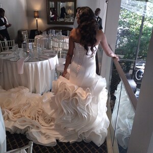 May include: A bride wearing a white wedding dress with a large, ruffled skirt. The dress has a fitted bodice and a lace-up back. The bride is standing on a balcony overlooking a garden.