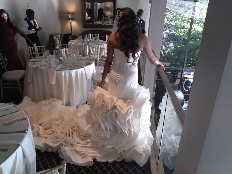 May include: A bride wearing a white wedding dress with a large, ruffled skirt. The dress has a fitted bodice and a lace-up back. The bride is standing on a balcony overlooking a garden.