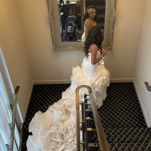 May include: A bride wearing a white wedding dress with a large, ruffled skirt. The dress is flowing down a staircase with black and gold patterned carpet.