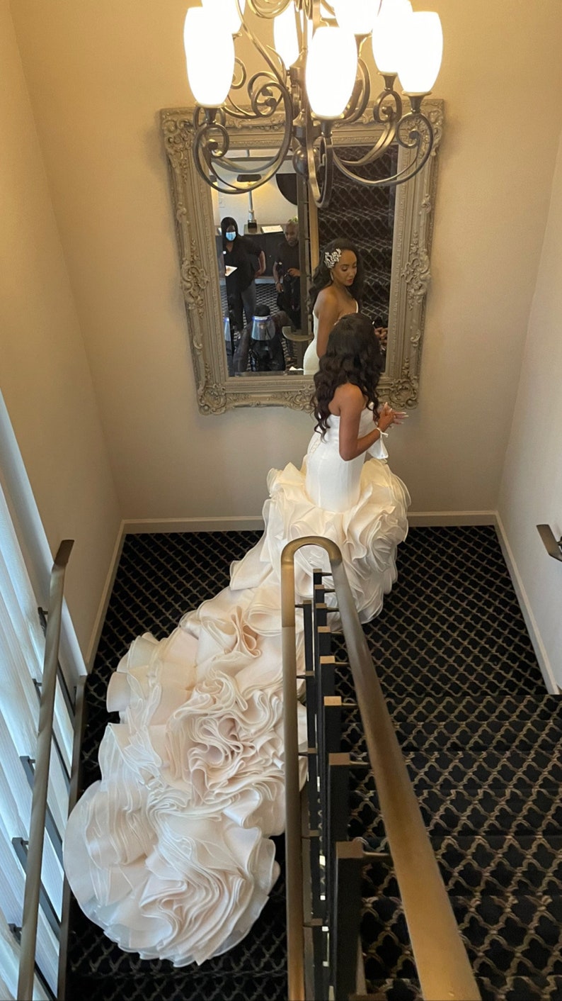 May include: A bride wearing a white wedding dress with a large, ruffled skirt. The dress is flowing down a staircase with black and gold patterned carpet.