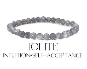 Puede incluir: Un brazalete de cuentas redondas de iolita gris. Las cuentas están ensartadas para formar una forma circular. La palabra "IOLITE" está impresa debajo del brazalete, junto con las palabras "INTUITION • SELF-ACCEPTANCE."