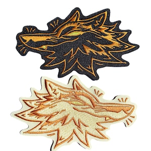 May include: Two embroidered patches featuring a stylised wolf head design. The top patch has a black background with metallic gold thread detailing. The bottom patch features a cream-coloured background with metallic gold thread detailing.