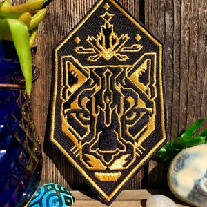May include: A black and gold embroidered patch with a geometric design of a panther's face. The patch is diamond-shaped and has a gold outline.
