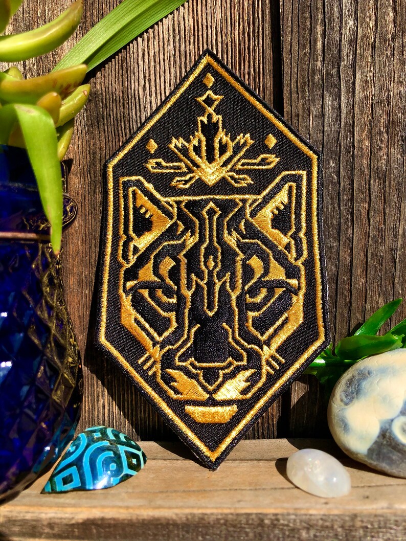 May include: A black and gold embroidered patch with a geometric design of a panther's face. The patch is diamond-shaped and has a gold outline.