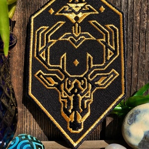 May include: A black and gold embroidered patch featuring a geometric deer head design. The deer head is surrounded by a hexagonal frame with a diamond pattern.