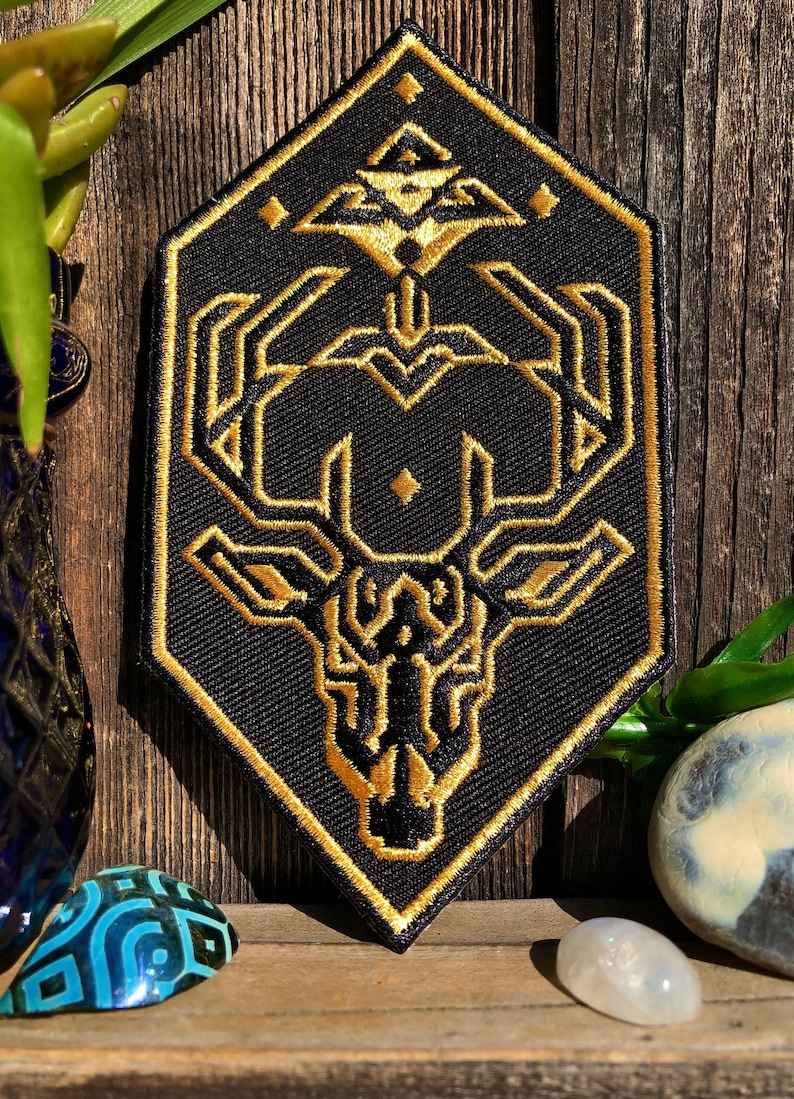 May include: A black and gold embroidered patch featuring a geometric deer head design. The deer head is surrounded by a hexagonal frame with a diamond pattern.