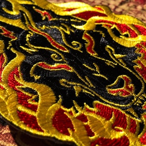 May include: Embroidered patch featuring a black dragon design with red and gold accents. The dragon's head is in the center, surrounded by flames. The patch is likely for clothing or accessories.