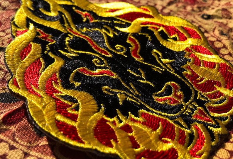 May include: Embroidered patch featuring a black dragon design with red and gold accents. The dragon's head is in the center, surrounded by flames. The patch is likely for clothing or accessories.