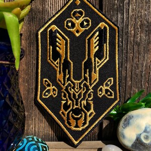 May include: A black and gold embroidered patch with a geometric design. The patch is shaped like a diamond and features a stylized creature with wings and a crown.