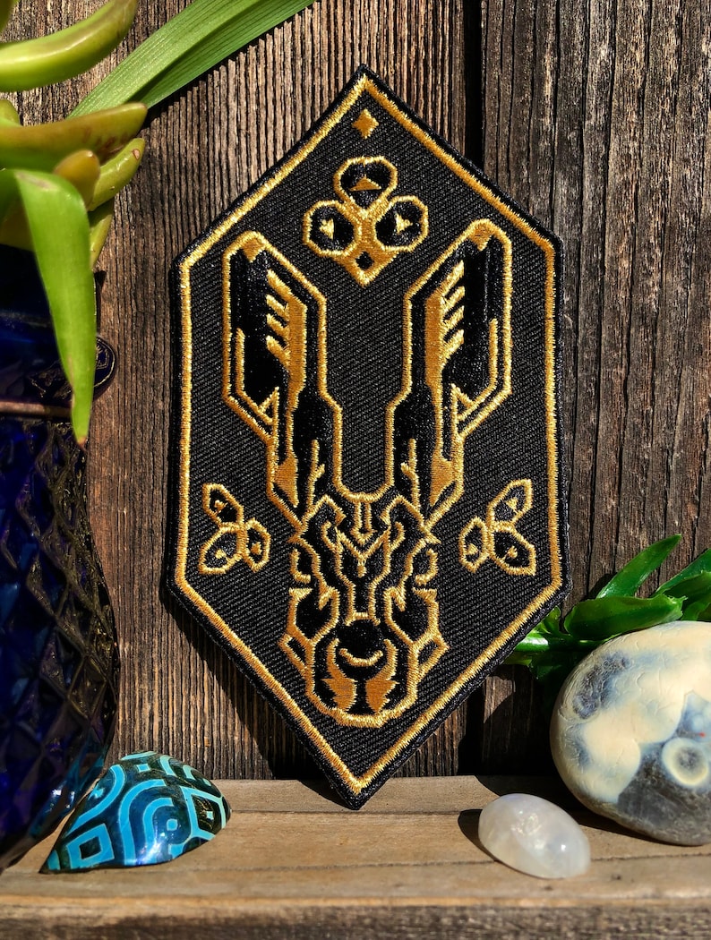 May include: A black and gold embroidered patch with a geometric design. The patch is shaped like a diamond and features a stylized creature with wings and a crown.