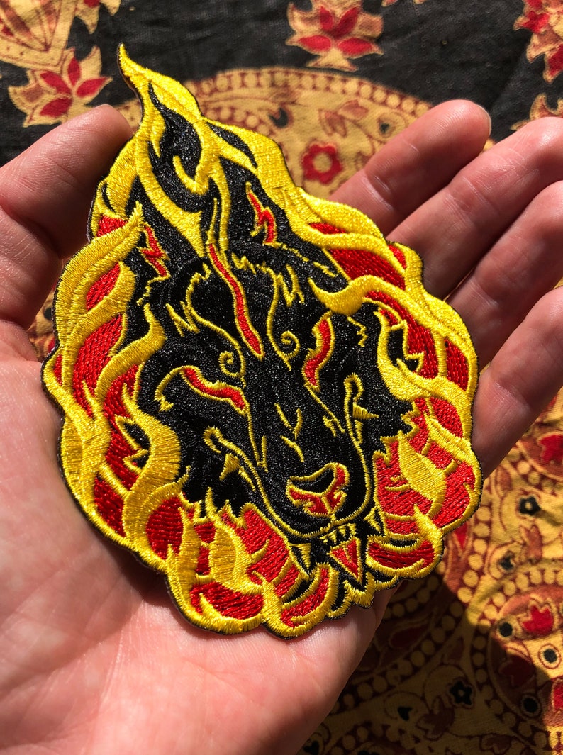 May include: Embroidered patch featuring a black wolf's head engulfed in flames. The flames are detailed with yellow and red thread, creating a striking contrast against the black. This patch is suitable for jackets, bags, or other fabric items.