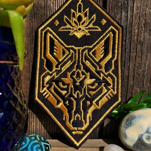 May include: A black and gold embroidered patch featuring a stylized wolf head with geometric patterns. The wolf's eyes are outlined in gold and the patch has a diamond shape.