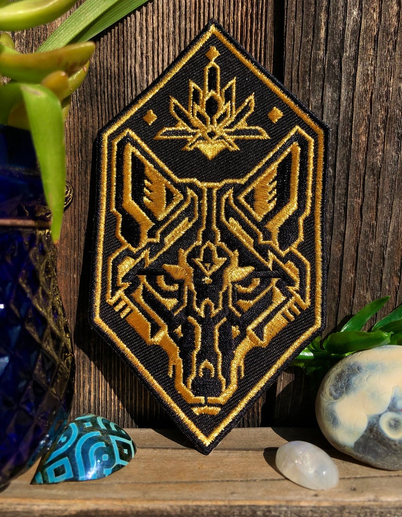 May include: A black and gold embroidered patch featuring a stylized wolf head with geometric patterns. The wolf's eyes are outlined in gold and the patch has a diamond shape.