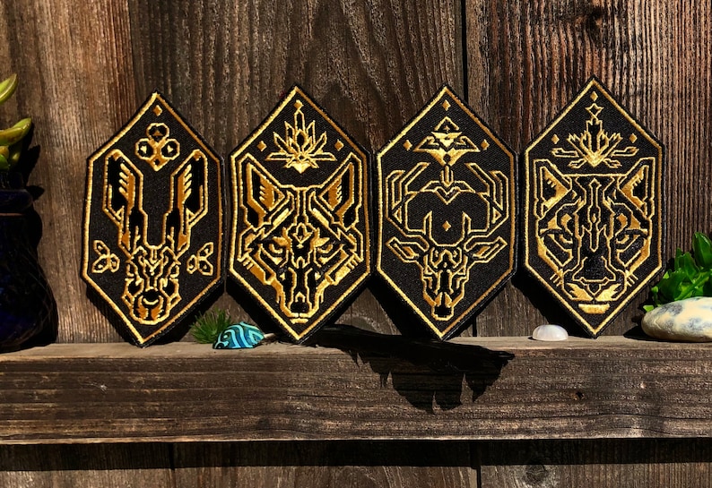 May include: Four black and gold embroidered patches featuring geometric designs of a wolf, a deer, a rabbit, and a cat. The patches are arranged in a row on a wooden surface.