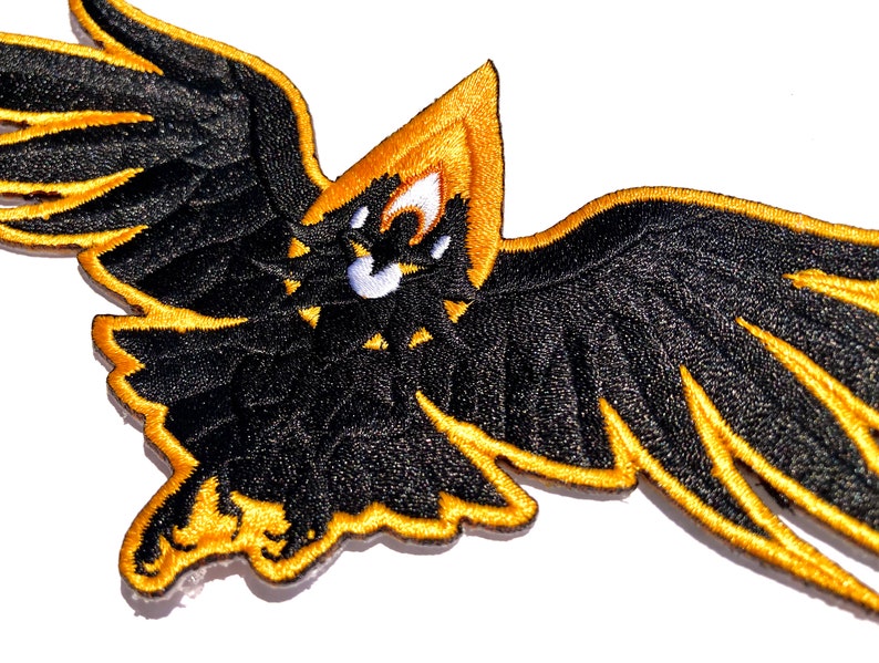 May include: Black and gold embroidered patch of a stylized eagle with its wings spread wide.