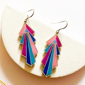 Puede incluir: Un par de pendientes de estilo Art Déco con un diseño geométrico. Los pendientes presentan una combinación de colores rosa, azul y morado con detalles metálicos plateados. Se exhiben sobre una superficie blanca semicircular.