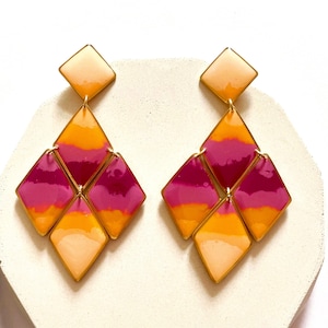 May include: A pair of geometric dangle earrings. Each earring features a square top and three diamond-shaped pendants in shades of peach, orange, and magenta, with gold-coloured metal accents. The earrings are displayed on a white surface.