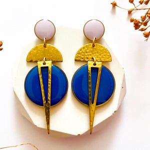 Puede incluir: Un par de pendientes geométricos con un diseño moderno. Cada pendiente presenta un círculo lila, un semicírculo dorado, un círculo azul y un triángulo dorado. Los pendientes se muestran sobre una superficie blanca.
