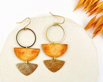 May include: A pair of gold earrings featuring three geometric shapes. The top shape is a circle, the middle shape is a semi-circle, and the bottom shape is a triangle. The semi-circle and triangle shapes are painted in a warm orange colour.