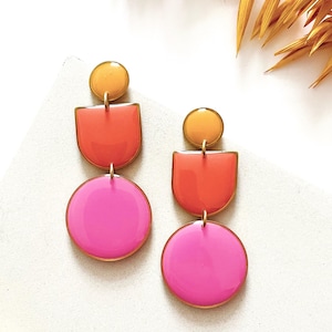 May include: A pair of vibrant, geometric earrings. Each earring features a small, round, yellow top piece, followed by a U-shaped orange section, and a large, round, pink bottom piece. The earrings have gold-colored edges and connecting hardware.