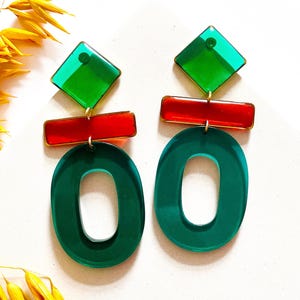 May include: Bold geometric earrings in green and red. The design includes green square studs, red rectangular bars, and large green oval drops. Gold-tone metal findings connect the elements. The earrings are displayed on a white surface.