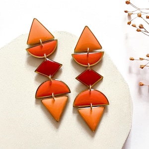 May include: Dangle earrings in geometric shapes, with orange, red, and gold colours. The earrings have a layered design with triangles, half-circles, and diamond shapes. The earrings are made of metal and enamel.