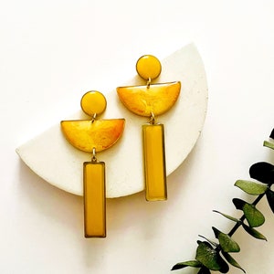 May include: A pair of yellow geometric dangle earrings with a semi-circle, circle, and rectangle shape. The earrings are made of resin and have a gold finish.