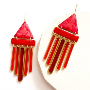 May include: A pair of red and gold geometric earrings. Each earring features a red triangle at the top, a red rectangle below, and several red vertical bars hanging down. The earrings have gold-colored metal accents and a hook for the ear.