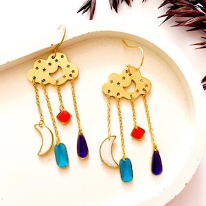 Puede incluir: Pendientes de nube dorados con recortes de estrellas y colgantes. Cada pendiente presenta un cuadrado rojo, un rectángulo azul, una lágrima morada y una luna creciente blanca. Los pendientes están sobre una bandeja blanca.