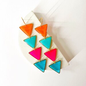 May include: Vibrant geometric earrings with a cascade of colourful triangles. The earrings feature orange, turquoise, and hot pink triangles, outlined in gold. The design is modern and eye-catching, ideal for adding a pop of colour to any outfit.